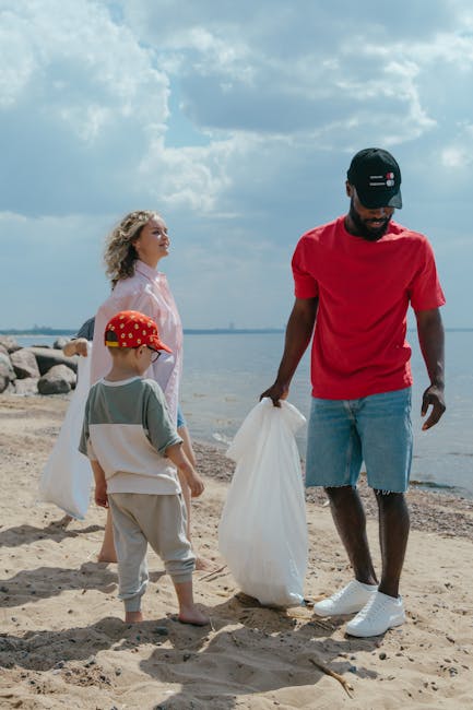 A group of three people, including a man and two children, are on a sandy beach near a body of water with a rocky shoreline in the background. The man, positioned on the right, is wearing a red t-shirt, light blue denim shorts, white sneakers, and a black cap, and is holding a large white plastic bag, possibly for rubbish collection. The young girl on the left has curly blonde hair, is dressed in a light pink blouse, and stands facing the water with her side to the camera. The boy in the center, slightly smaller, is wearing a grey t-shirt and beige trousers, with a red cap decorated with yellow and white spots; he is holding another white plastic bag, likely involved in an environmental activity or positive waste management effort. The scene is illuminated by natural daylight, with a partly cloudy sky overhead, and the overall atmosphere suggests an outdoor cleanup activity, potentially related to environmentally conscious rubbish removal or local waste management practices in an outdoor setting. The image aligns with themes of private disposal and on-site clearance, highlighting environmentally responsible waste handling near a natural waterway, consistent with the services of Rubbish Removal Barnet.