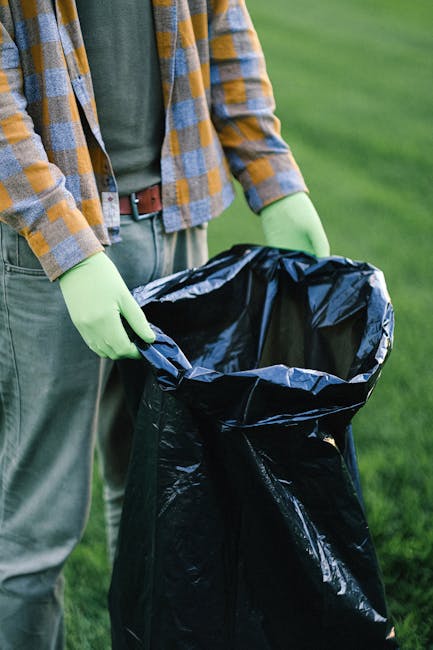 A person wearing a white T-shirt with the text 'NO PLANT B' printed in green on the front, is holding a clear plastic bag filled with crumpled disposable masks and gloves, which appear to be made of synthetic material. The individual is wearing white gloves to handle the waste, and a black wristwatch is visible on their left wrist. The background shows an outdoor setting with blurred natural elements, such as grass and trees, illuminated by warm natural sunlight, suggesting a late afternoon or early evening. The scene reflects responsible handling of potentially contaminated waste, with contextual relevance to rubbish removal and waste management services, emphasizing proper disposal practices that are vital in private or alternative waste collection scenarios, like those provided by Rubbish Removal Barnet.
