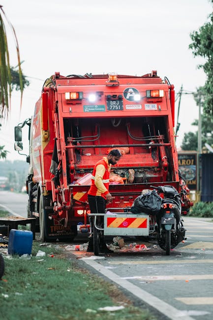 A waste collection worker wearing a red safety vest is standing beside the rear opening of a large red rubbish truck parked on the side of a paved street. The truck’s rear compactor mechanism is visible, showing a textured metal surface with some debris and waste materials inside. The worker appears to be in the process of managing or loading waste, with a black plastic bag placed on a small metal platform attached to the truck. The scene includes a patch of grass and sidewalk to the left, with some small scattered waste and a blue bin nearby. In the background, there is a tree, utility poles, and a building with a sign, indicating an urban or suburban environment. The overcast sky and diffused lighting suggest daytime, and this setting aligns with on-site waste collection services by companies like Rubbish Removal Barnet, supporting private waste handling outside of municipal collection. The scene emphasizes the practical operation of a rubbish removal service focusing on residential or commercial waste disposal in a street context.