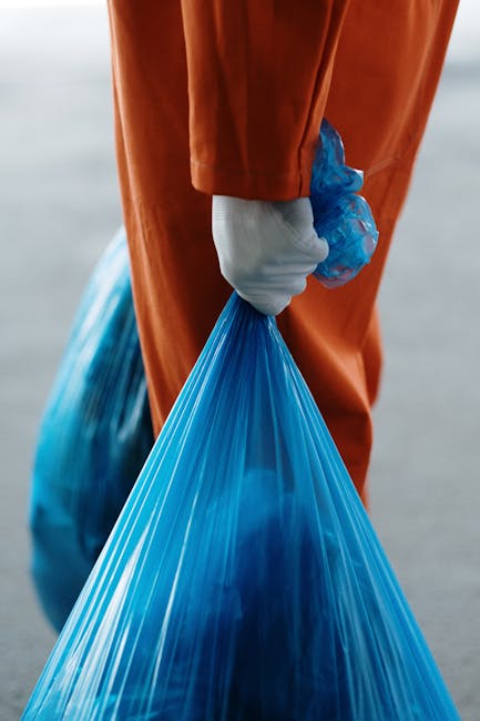 A person dressed in orange protective overalls and white footwear is standing on a plain grey floor, holding two large transparent blue plastic rubbish bags filled with waste material, one in each hand. The individual’s upper body and head are not visible in the image. In the foreground, part of an orange and black portable sprayer or cleaning equipment with a flexible hose is partially visible, resting on the ground. The background features a smooth, light grey wall, creating a neutral environment suitable for showcasing rubbish collection or waste removal activities. The scene emphasizes the handling of bulk waste, aligning with professional rubbish removal services offered by companies such as Rubbish Removal Barnet, which specializes in convenient, on-site clearance for both residential and commercial clients.