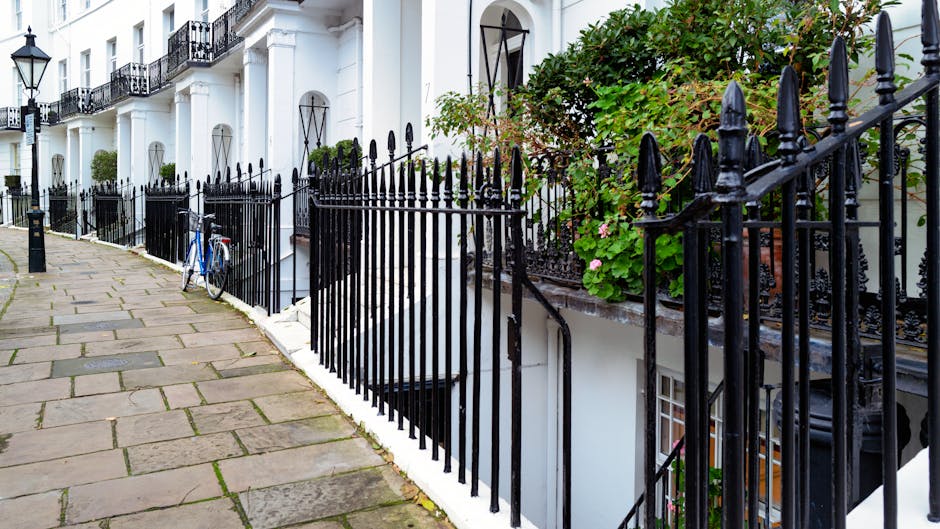 A row of white terraced houses with black wrought iron fences enclosing small front gardens and steps leading down to basement entrances. The fences feature pointed finials and decorative elements. On the pavement in front of the houses, there is a blue bicycle leaning against the fence, with a black bollard positioned nearby. The front gardens contain green leafy plants and flowering shrubs, some with pink blossoms, adding natural contrast to the black fences and white facades. The scene is illuminated by natural daylight, highlighting the textures of the stone-paved sidewalk, the smooth white building surfaces, and the glossy finish of the fences. The image appears to depict a quiet residential street, where private waste disposal may be handled independently, supported by local rubbish removal services such as Rubbish Removal Barnet, evident through the awareness of on-site clearance methods fitting with the context of alternative rubbish handling for terraces.