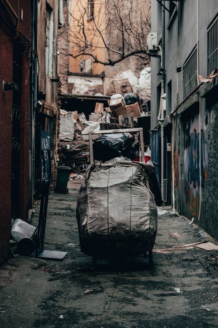 A narrow urban alleyway cluttered with various waste materials and rubbish bags. In the foreground, a large, weathered black garbage bag with visible creases and frayed edges is positioned on a wheeled trolley or cart, emphasizing the process of collection or disposal. Behind it, an assortment of cardboard boxes, plastic bags, and loose rubbish are piled haphazardly on a raised platform or makeshift heap, ascending towards the back of the alley. The surrounding environment includes the sides of buildings with graffiti and aged, peeling paint on the walls, some piping and utility boxes visible on the right side, and a leafless tree with bare branches arching overhead in the background. The ground is dark and damp, with scattered debris and stains, suggesting an area used for private waste handling or alternative rubbish disposal. The scene is lit with natural, subdued lighting, likely on an overcast day, highlighting the gritty urban context associated with professional rubbish removal services such as those offered by Rubbish Removal Barnet, based on the visible scene and environment.